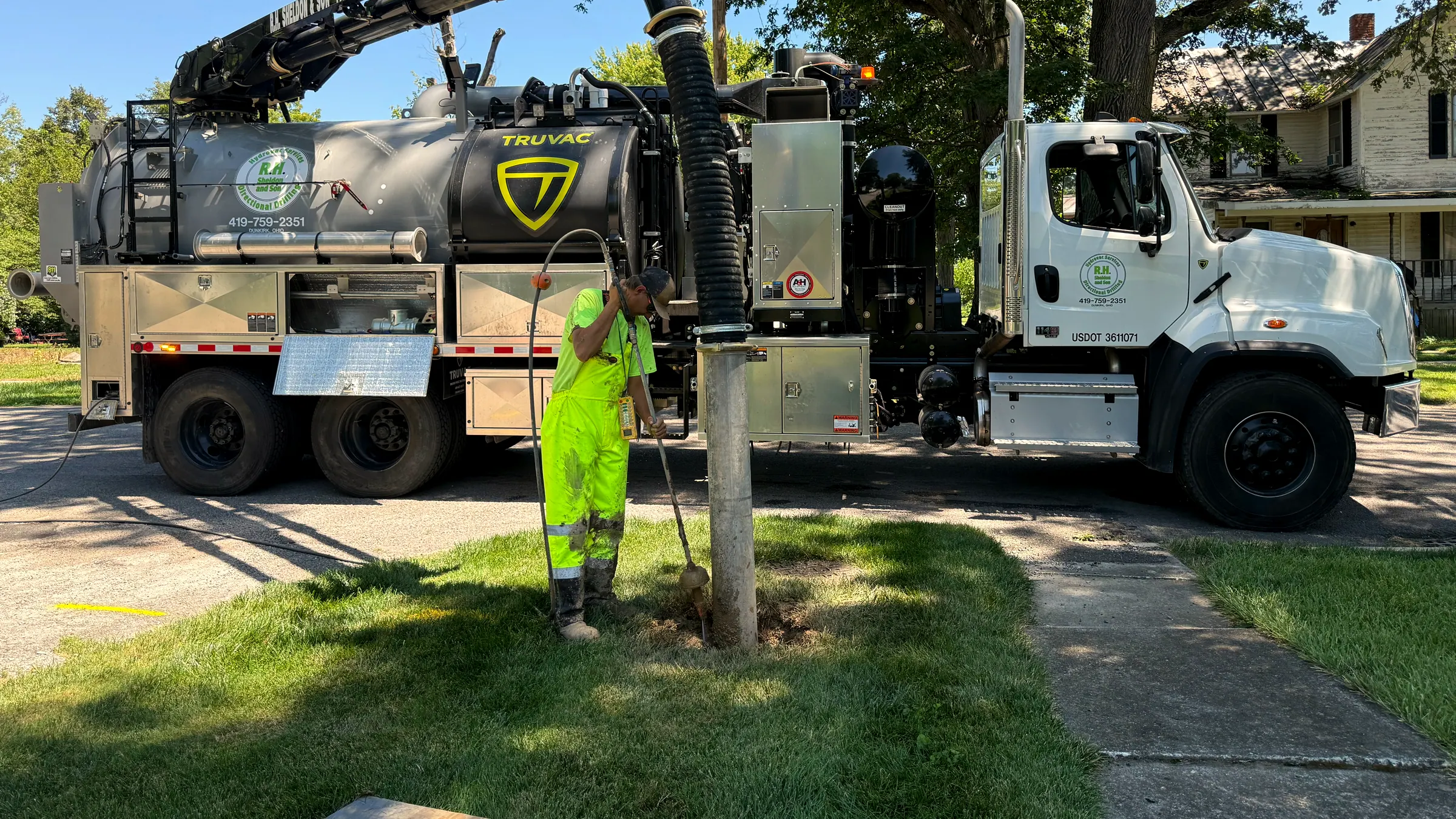 An R.H. Sheldon & Son crew member in full hi-vis potholing by hand-held wand beside a utility pole, with the vac truck parked on the street behind him.